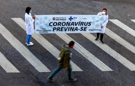 Duas mulheres estão em cima da faixa de pedestre segurando uma faixa que diz "Coronavírus previna-se."